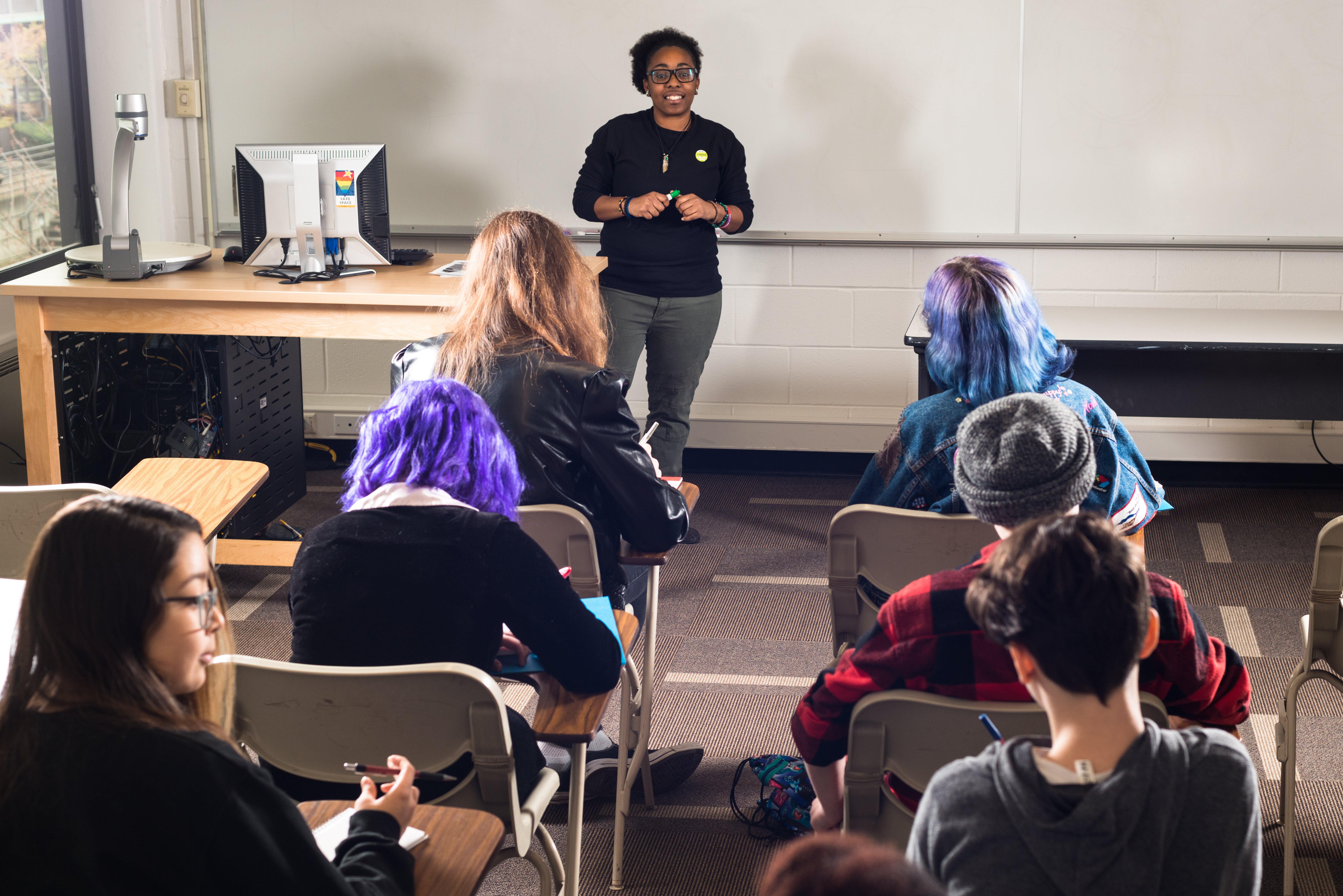 An educator standing in front of a whiteboard teaches a classroom of six students at their desks.